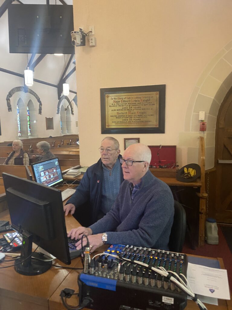 Two older men work at a sound mixing console and computer in a church office area with pews in view.